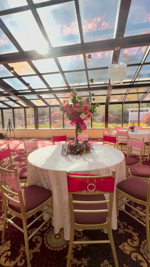 Round reception table dressed in cream with red chair sashes and a tall pink centrepiece, under a glass roof.