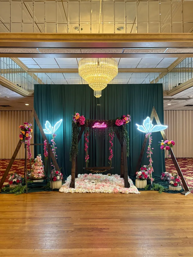 Dark green draped backdrop lit with neon butterflies, flanked by floral arches and a petal-strewn floor.